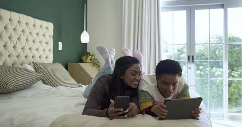 Diverse Female Friends Relaxing with Gadgets on Bed in Bright Bedroom