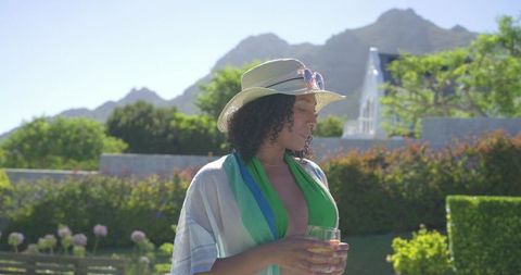 African american woman enjoying drink in sunlit resort garden with straw hat and swimsuit