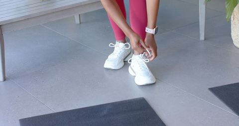 Tying white sneakers before workout wearing pink leggings and smartwatch at home gym