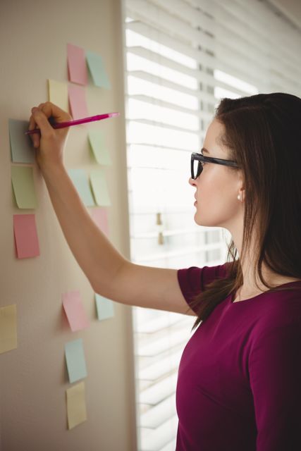 Woman writing on colorful sticky notes on wall by window, wearing glasses and casual clothing. Ideal for themes related to productivity, planning, brainstorming, and home office setups. Can be used in articles, blogs, or advertisements focusing on organization, creativity, and efficient work habits.
