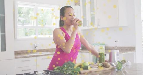 Korean woman drinking green smoothie in sunlit kitchen making healthy juice for wellness