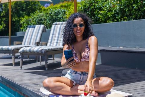Smiling african woman relaxing poolside in zebra top and denim shorts holding smartphone