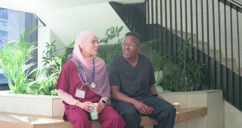Diverse medical team taking coffee break on bench in bright modern hospital lobby with greenery