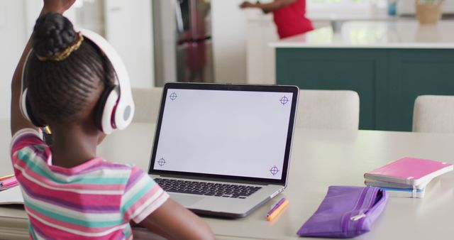 Young African American girl engaging in an online class from home. She raises her hand while facing an open laptop with headphones on. Books and writing materials are neatly arranged on the table. Background includes a kitchen, indicating a home environment. Ideal for use in articles, blogs, and resources that discuss distance learning, online education, and the integration of technology in home schooling.