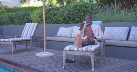 Mature african american man relaxing on poolside lounger eating snack by resort pool deck