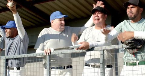 Baseball teammates cheering in dugout leaning on rail holding bat and glove celebrating