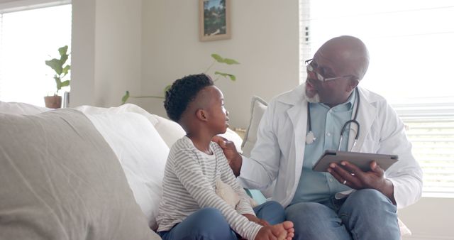 Doctor with stethoscope reassuring boy during home consultation while holding a digital tablet. Ideal for healthcare services promotional materials, pediatric health articles, and family medical care resources.