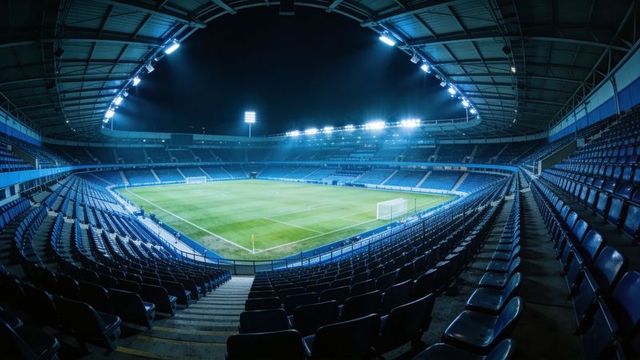 Floodlit football stadium interior at night featuring covered roof and empty seating