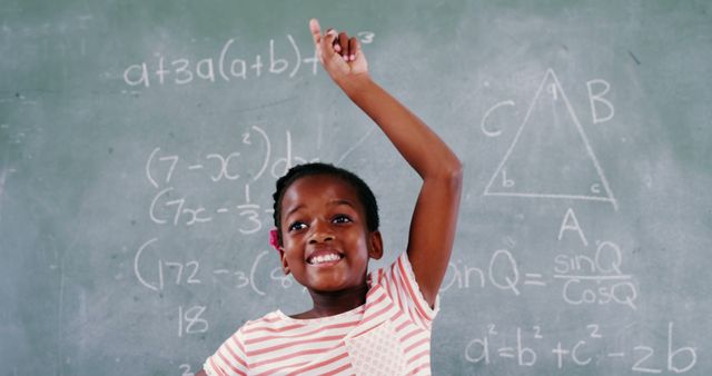 A young African American girl stands in front of a chalkboard filled with mathematical equations, raising her hand with confidence. Her bright smile and raised hand suggest enthusiasm and readiness to answer a question in a classroom setting.