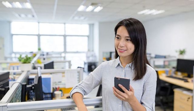 Asian professional woman holding smartphone and leaning on cubicle in open-plan office