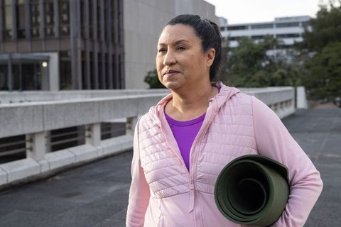 Mature woman walking with yoga mat on urban walkway wearing pink hoodie for wellness
