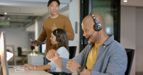 Diverse team collaborating in open office wearing headsets with tablet celebrating success