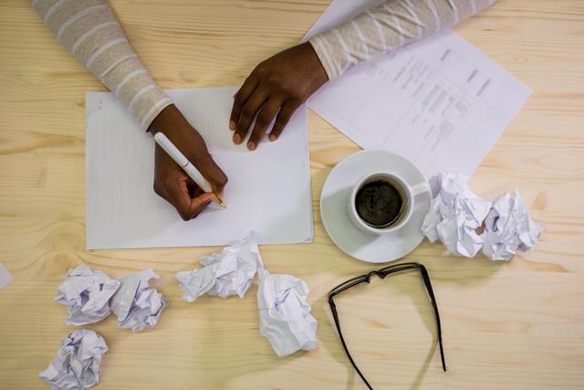 Hands of a woman writing on blank paper at a wooden desk, surrounded by crumpled notes, a cup of coffee, and glasses. Ideal for illustrating concepts of brainstorming, creativity, work, and productivity. Suitable for use in articles, blogs, and advertisements related to writing, office work, and creative processes.