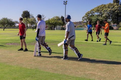 Youth cricketers walking across sunlit park field carrying bats and pads during practice session