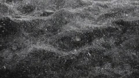 Macro video of fine particles settling on rippled dark surface, grayscale speckled texture
