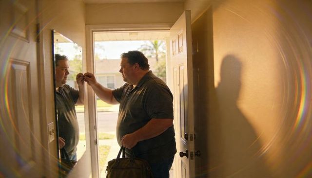 Man pausing at sunlit front door holding keys and duffel bag with mirror reflection