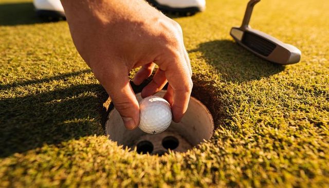 Golfer placing ball into cup on sunlit putting green with putter closeup