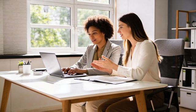 Two female colleagues collaborating on laptop in bright modern office discussing documents