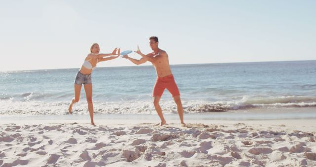 A young Caucasian woman and man are playing with a frisbee on a sunny beach, with copy space. Their active lifestyle and enjoyment of beach sports are evident in their playful interaction.