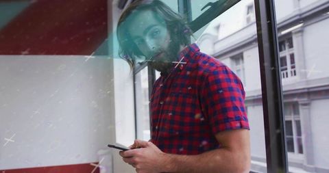 Bearded man in red plaid shirt scrolling smartphone by office window with reflections