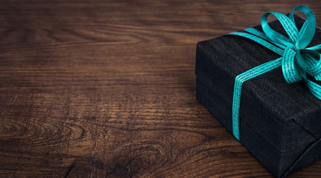 Beautifully wrapped gift box with a teal ribbon sitting on a brown wooden surface, perfect for use in advertisements, social media posts, or websites related to celebrations, gifts, and luxury items. Suitable for illustrating concepts of gifting, special occasions like birthdays and anniversaries, and creating a festive mood.