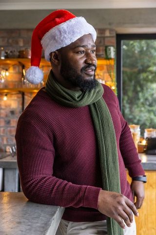 African american man wearing santa hat and scarf leaning on kitchen countertop, festive holiday