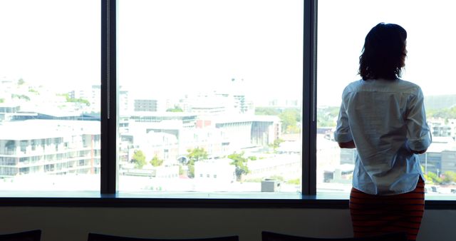 Businesswoman standing by office window, overlooking cityscape. Ideal for topics on career, urban professional life, business environment, contemplative moments in workplace, and modern office settings.