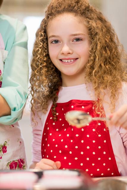 Young girl with curly hair wearing a red polka dot apron, smiling while baking cupcakes in a home kitchen. Ideal for use in content related to family activities, childhood learning, cooking tutorials, and home baking.