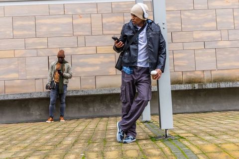 Leaning commuter checking smartphone and holding coffee near tiled wall in urban plaza