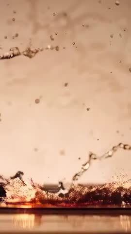Vertical video capturing amber liquid splashing, droplet crown and backlit reflections
