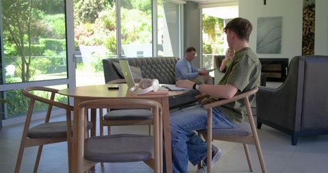 Teenager working on laptop at wooden dining table in sunlit modern living room
