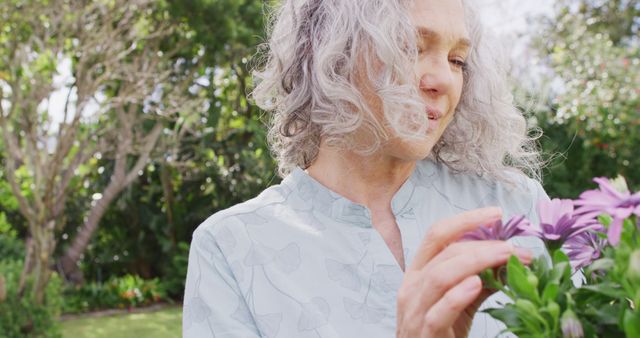 An elderly woman with grey hair stands in a lush green garden, gently tending to flowers. This can be used in lifestyle, gardening, or nature-related content, promoting leisure, horticulture, and outdoor activities for seniors.