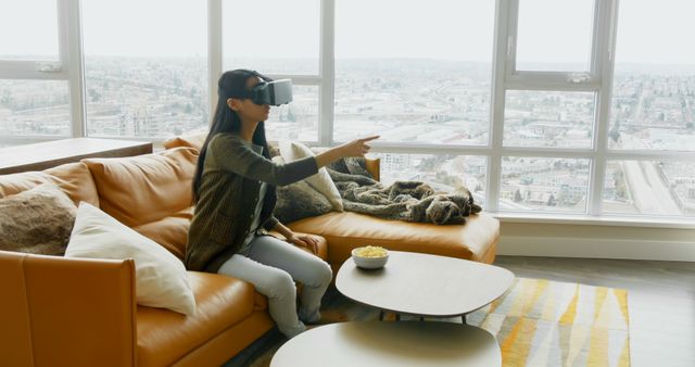 Young woman sitting on orange couch wearing VR headset, pointing at virtual object. Comfortable modern living room with city view through large windows. Ideal for technology, modern lifestyle, and entertainment concepts.