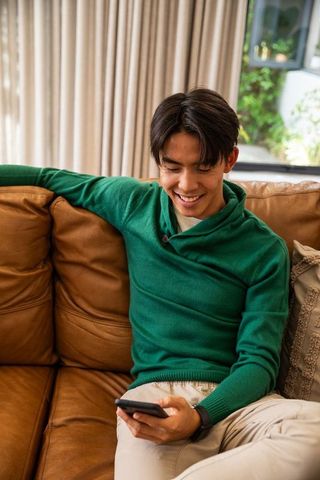 Smiling asian young man relaxing on brown leather sofa using smartphone for messaging