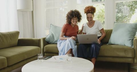 Diverse women collaborating on laptop and charts while smiling on living room sofa home workspace