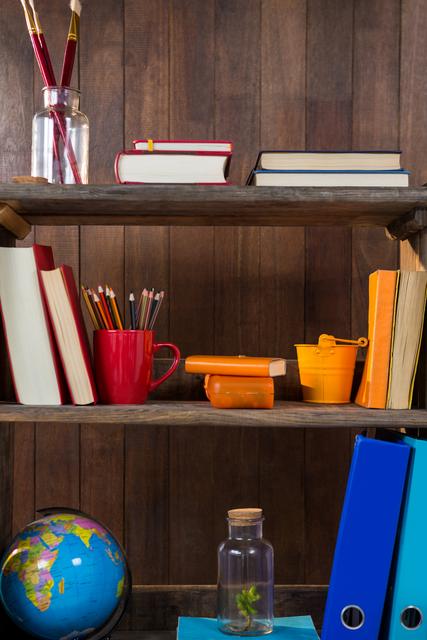 Books and stationery items are neatly arranged on a wooden shelf. The shelf includes books, pencils in a red mug, a small globe, a jar, and colorful binders. This image can be used for educational content, office organization tips, or interior design inspiration.