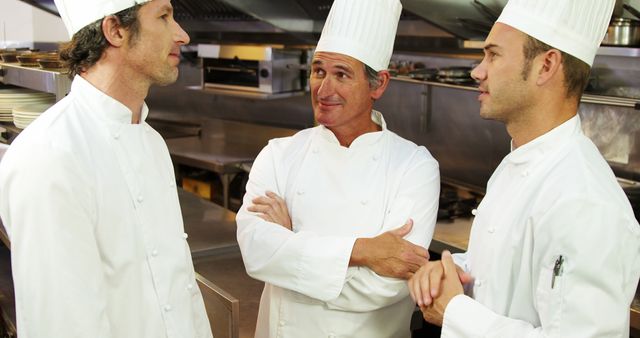 Three chefs in traditional white uniforms are standing in a professional kitchen discussing recipe ideas. They appear engaged in conversation, indicating teamwork and collaboration. This image can be used in presentations related to culinary arts, restaurant management, cooking classes, teamwork in restaurants, and kitchen professionalism.