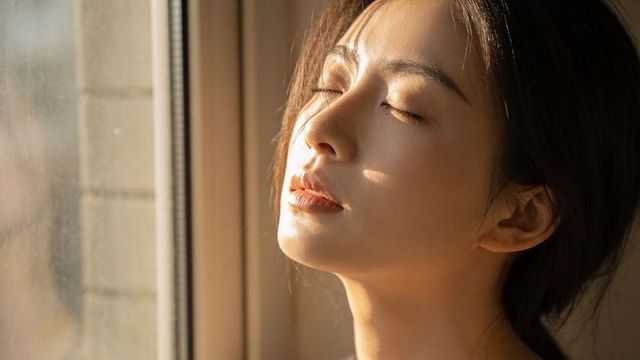 Asian woman basking in golden window sunlight with closed eyes, serene intimate portrait