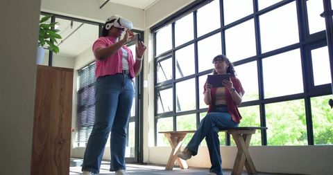 Female colleagues testing vr headset and using tablet in sunlit modern tech workspace