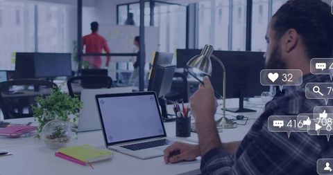Bearded man typing on laptop and smartphone in open-plan office with social media overlays