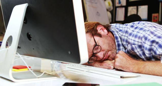 A tired man with glasses is resting his head on his hands at his desk with a computer in an office environment. This image can be used to depict workplace stress, fatigue, burnout, or the importance of taking breaks in a professional setting. Ideal for articles, blogs, advertisements, or posts about work-life balance, mental health, and productivity.