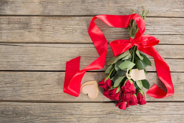 This image shows a bunch of red roses tied with a red ribbon, accompanied by a heart-shaped decoration on a wooden background. Ideal for use in romantic contexts such as Valentine's Day, anniversaries, or special occasions. Perfect for greeting cards, social media posts, or advertisements promoting love and romance.