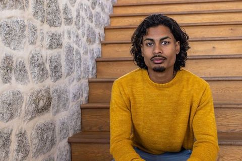 Indian man sitting on wooden staircase wearing mustard sweater, thoughtful indoor portrait