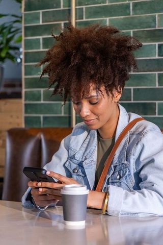 African american woman texting at cafe wearing denim jacket holding smartphone and coffee