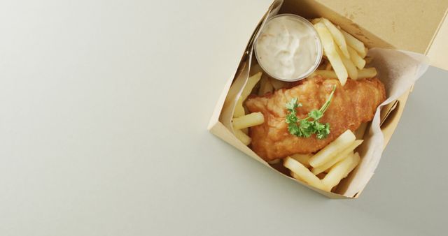 A takeout box containing golden fried fish and crispy chips with a small cup of tartar sauce on the side, garnished with fresh parsley. Ideal for depicting traditional British fast food, takeout meals, and comfort food. Suitable for use in food blogs, menus, and advertisements.