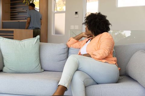 African american woman sitting on sofa looking away, partner walking in sunlit living room