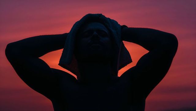 Silhouetted shirtless man holding towel behind head at sunset with glowing sun halo