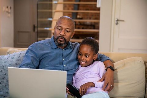 African american father sharing laptop with daughter on couch, smiling and learning