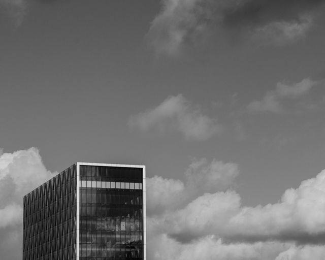 Black and white photograph of a modern office building with a cloudy sky background. Suitable for business materials, architectural portfolios, and presentations. Can also be used to convey modernity and urban lifestyle in advertisements and editorial pieces.