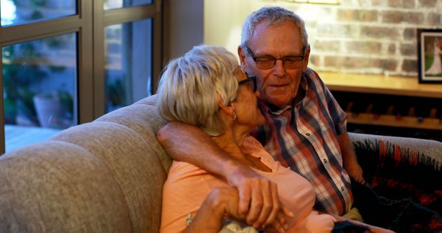 A senior Caucasian couple shares a tender moment together on a couch, with warm lighting adding to the cozy atmosphere. Their affectionate embrace reflects a deep bond and years of companionship.